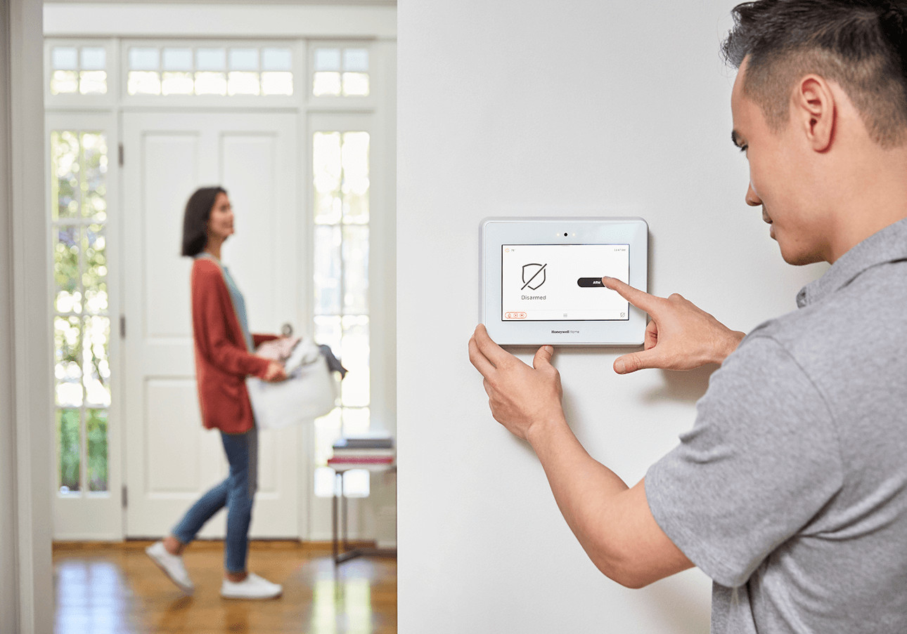 RESIDENTIALSERVICES-SECURITYANDFIRE Person adjusts settings on a wall-mounted tablet while another person, carrying a bag, walks in the background of a bright room with a front door.