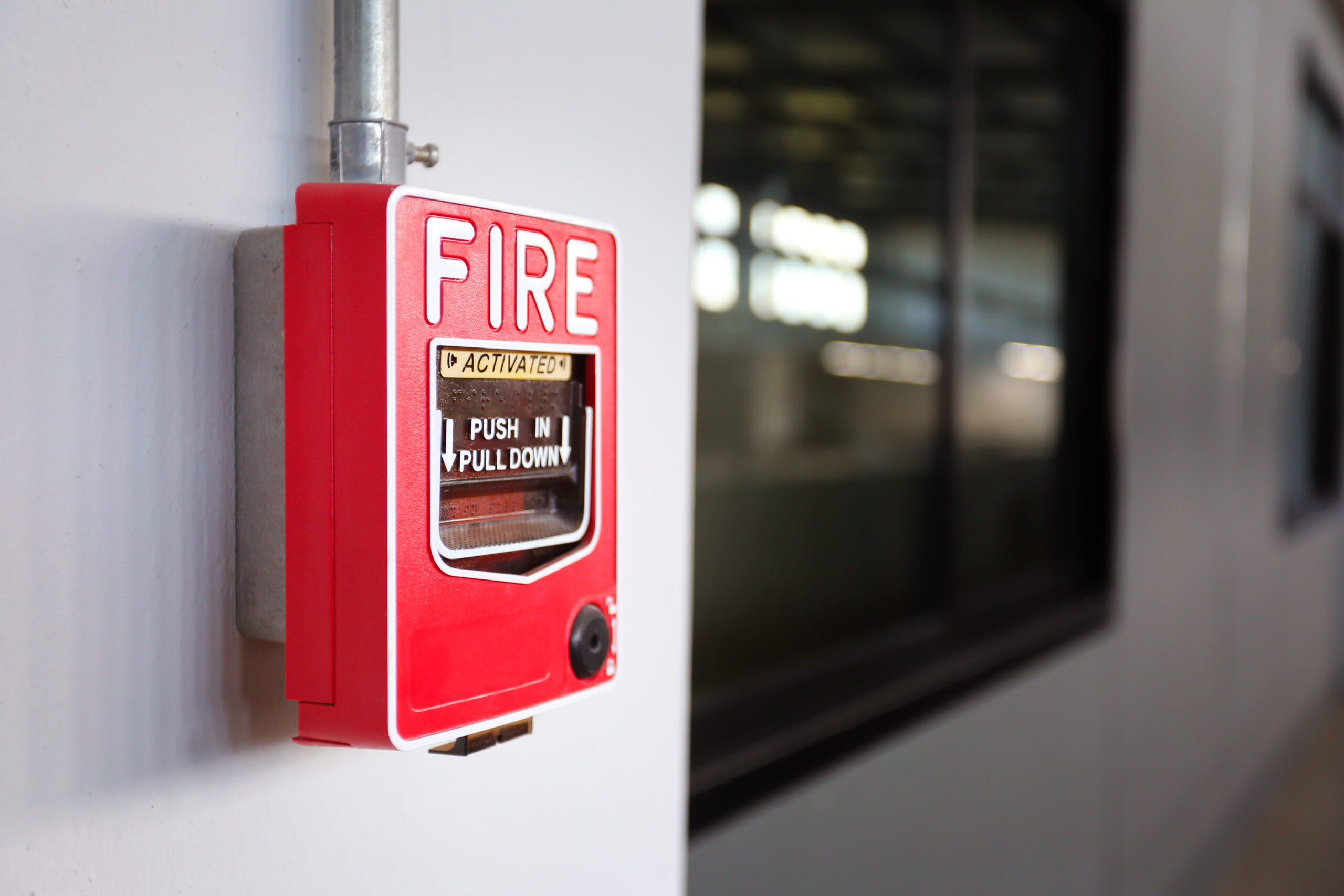 Fire,Alarm,Switch,On,The,Factory,Wall. Red fire alarm box mounted on a white wall near a window, with instructions to "push in pull down" visible on the front.
